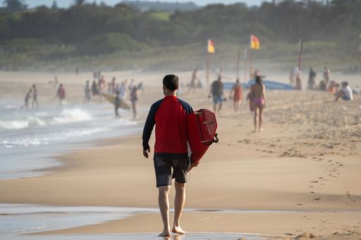 A shallow focus of a surfer walking on the shore with a surfboard under his arm.