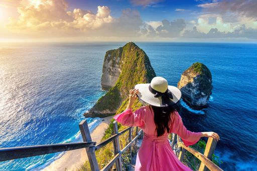 An aerial view of a woman in a hat and dress standing over the Kelingking Beach in Bali, Indonesia