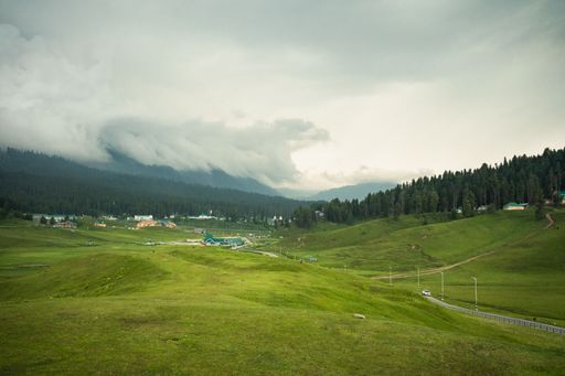 A shot of a green grass field surrounded by large trees and hills on a cloudy day
