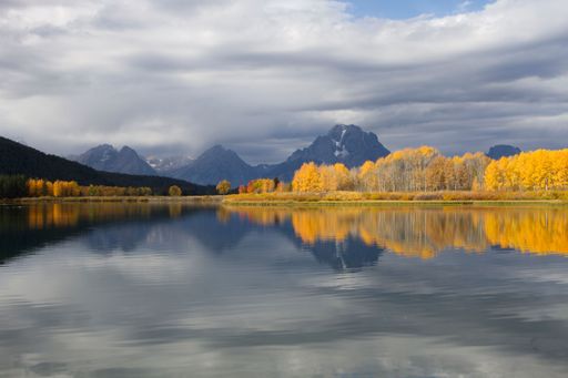 The view of yellow trees and mountains reflecting in the lake under the cloudy sky
