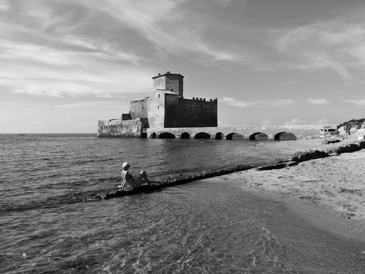 A girl with a hat sitting on a rock looking at the medieval tower, grayscale shot