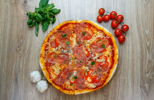 A top view of pizza with prosciutto Crudo, mushrooms, mozzarella, red bell peppers, parsley with ingredients next to it on a wooden table