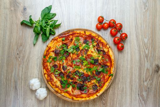 A top view tasty pizza with sausage, ham, salami, tomato sauce and parsley on a wooden table with greens, cherry tomatoes and garlic beside