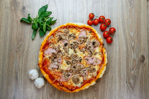 A top view of Pizza capricciosa with cherry tomatoes,greens and garlic next to it on a wooden table
