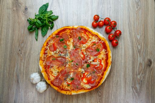 A top view of pizza with prosciutto Crudo, mushrooms, mozzarella, red bell peppers, parsley with ingredients next to it on a wooden table