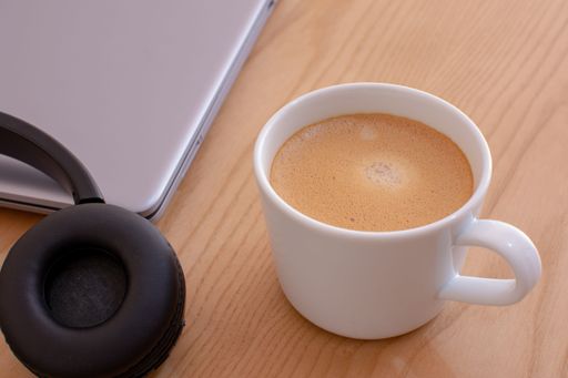 A closeup shot of a cup of coffee with headphones and laptop on the side on wooden table