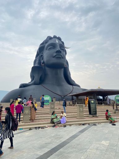 A vertical shot of people sitting on stairs against the Adi yogi statue on a cloudy day
