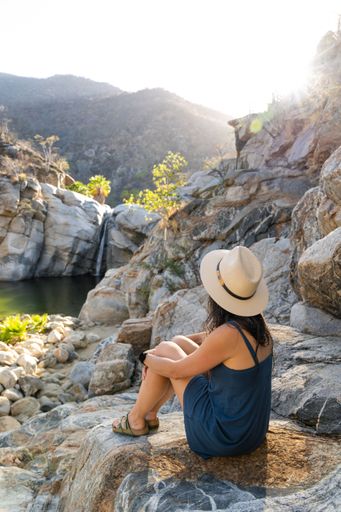 A vertical shot of a woman sitting on a rock and enjoying the view over a small water pond and a waterfall with hills in the background