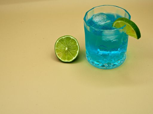 A blue cocktail served with a slice of lime on a neutral yellow background