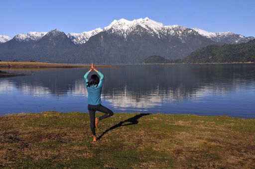 A caucasian woman wearing sportswear, holding a yoga pose in front of the calm lake and beautiful mountains during daylight