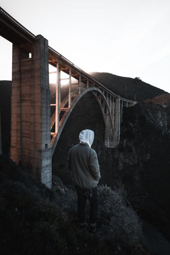 A vertical shot of a guy standing near a bridge on the edge of a cliff