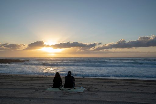 A back view of a couple sitting on the sandy beach watching the sea during cloudy sunset