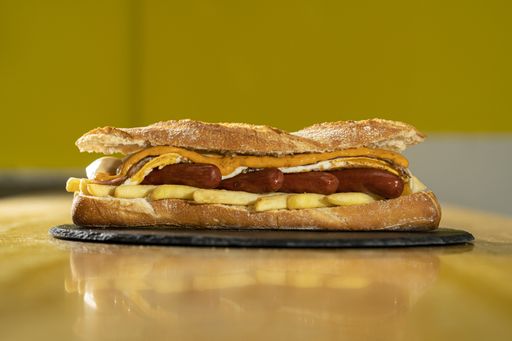 A closeup shot of a sandwich with baguette, sausages, and french fries on a black round board on a wooden table
