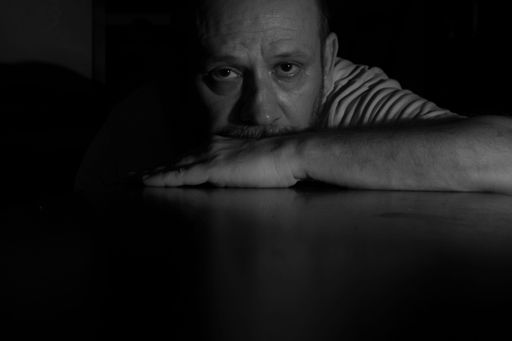 A grayscale closeup of a man in the darkness leaning on the table and looking at the camera.