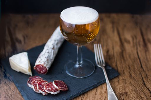 A closeup shot of a glass of beer with brie cheese and smoked sausage on a board