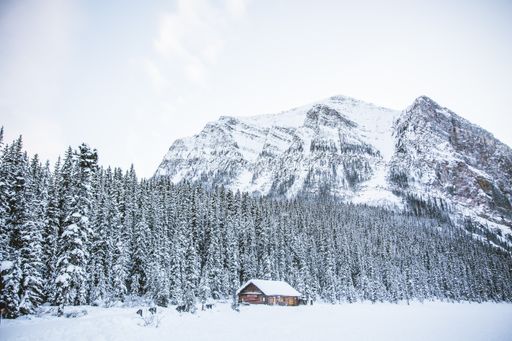 A hut in a snowy field with rocky mountains and a thick forest