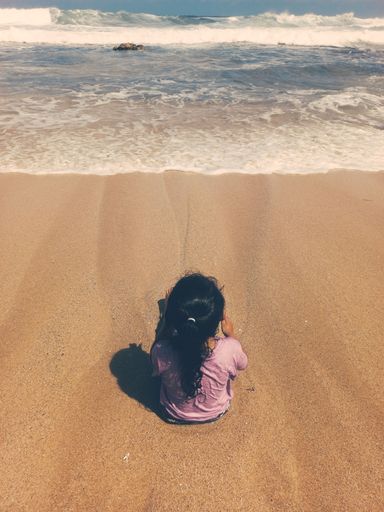 A vertical shot of a little girl sitting on the beach against a tide