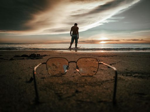 A closeup of glasses on the beach with a standing man in the background of the sunset.