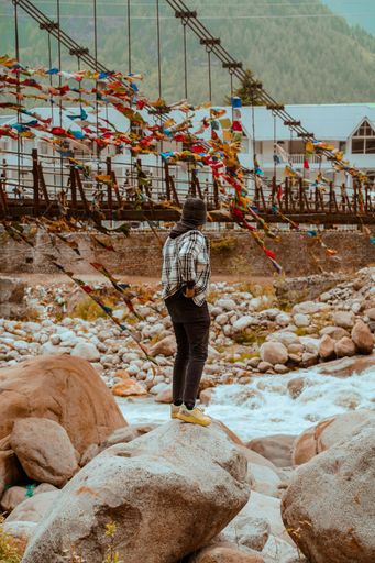 A vertical shot of a young man standing on a rock and looking at the footbridge. Manali, India.
