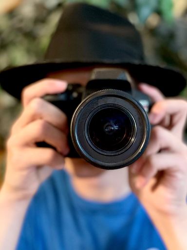 Young man in black fedora hat and blue shirt holding camera in front of a tree. Blurred backgroun