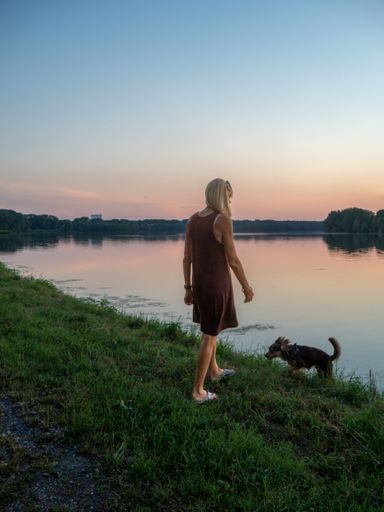 A vertical of a blonde woman wearing a casual dress enjoying the sunset at river bank with a dog