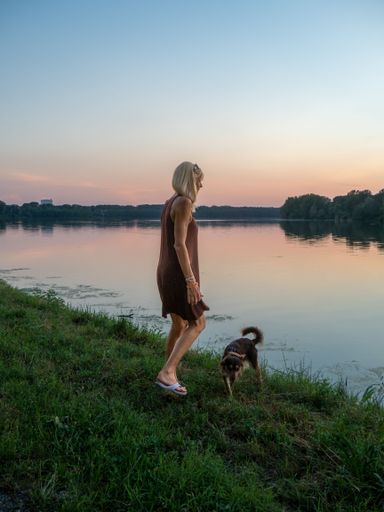 A vertical of a blonde woman wearing a casual dress enjoying the sunset at river bank with a dog
