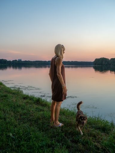 A vertical of a blonde woman wearing a casual dress enjoying the sunset at river bank with a dog
