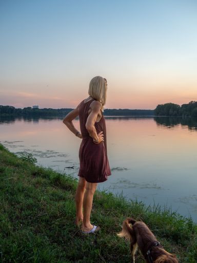 A vertical of a blonde woman wearing a casual dress enjoying the sunset at river bank with a dog