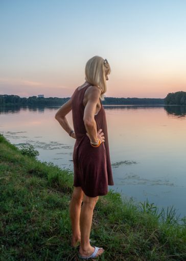 A vertical of a blonde woman in a casual dress enjoying the sunset by a river with hands on her hips