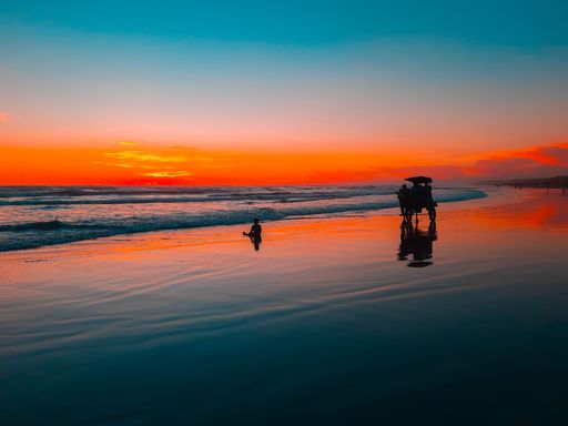 a horse-drawn carriage with a group of people driving at sunset time on the wet and reflecting sand of Parangtritis Beach, Java, Indonesia