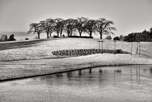 A grayscale shot of fields partly covered in snow by a pond