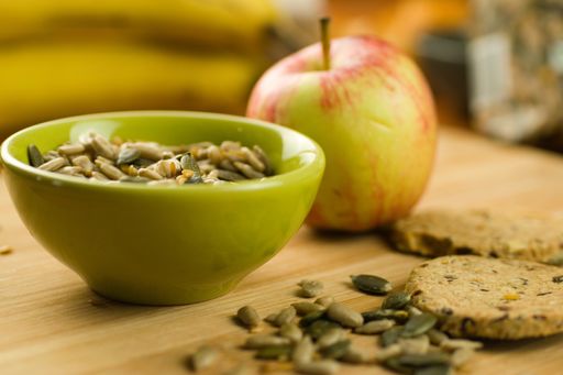 A closeup shot of sunflower and pumpkin seeds in a bowl, an apple, and oatcakes