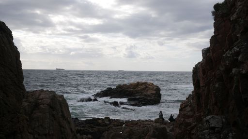 A scenic view of people sitting on rocks watching ocean waves crashing against beach on a gloomy day