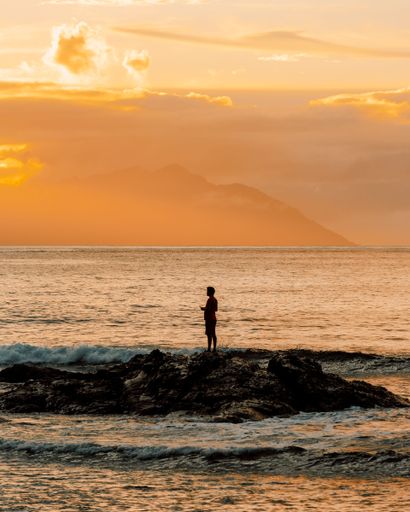 A vertical shot of a gorgeous sunset at sea with the silhouette of a man standing on rocks under a golden sky