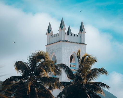 A white castle tower with spiky blue roofs surrounded by palm trees under a bright cloudy sky
