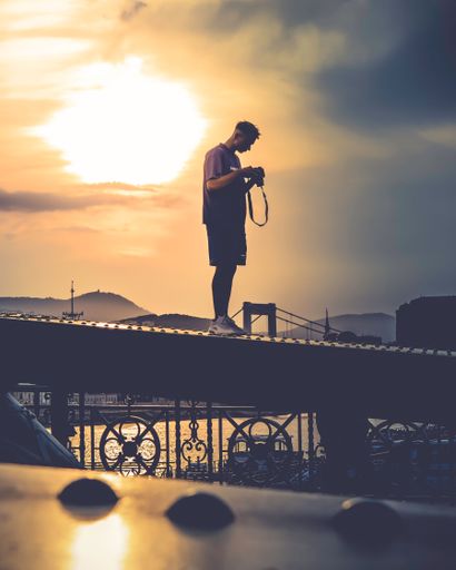 A vertical shot of a Caucasian guy looking at his camera, standing on a bridge at a moody sunset