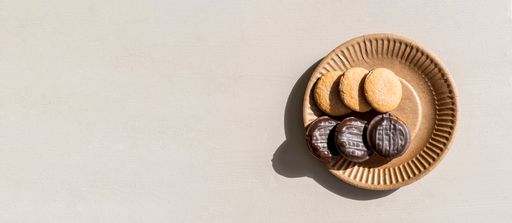 On light surface of kitchen table is a brown paper plate with delicious chocolate chip cookies. Negative space on left.