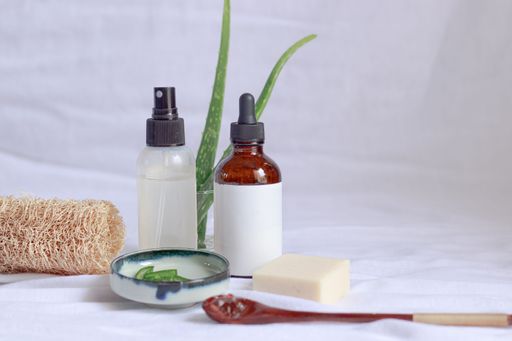 A closeup of bottles filled with liquid around aloe leaves and a spoon on a white cloth