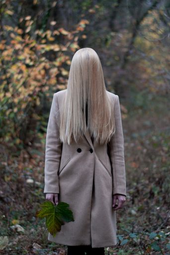 A vertical shot of a woman standing in a forest in a camelia coat with long blonde hair covering her face and a big leaf in her hands