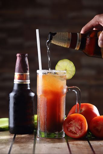 A hand pouring beer into a glass of iced cocktail with tomato and lime
