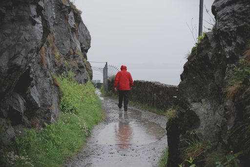 Back view of a man in wet red jacket who walks in the rain o the trail of the Bray to Greystones cliff walk. Rainy and foggy Irish weather.
