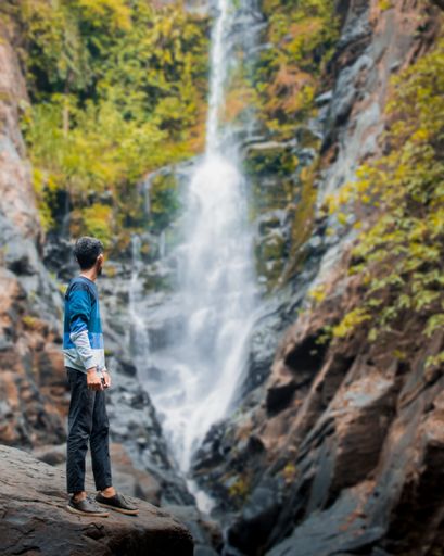 A vertical back shot of a young male standing on a rock looking at blurry Savri waterfall