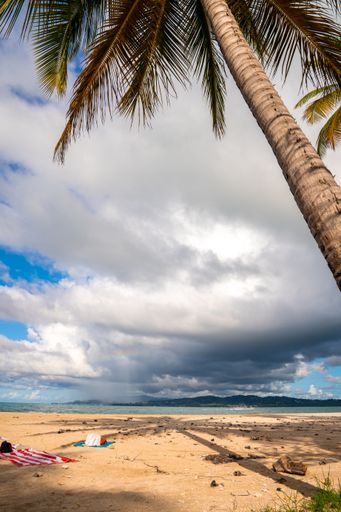 A vertical shot of a beach surrounded by the sea and hills under a blue cloudy sky and sunlight