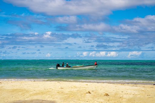 A beach surrounded by the sea with people in a boat on it under the sunlight and a blue cloudy sky