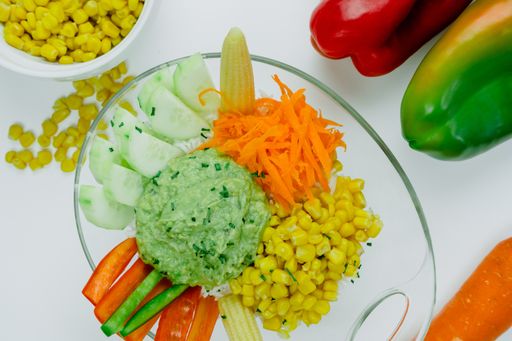 A top view of a bowl with sliced carrots, cucumbers, peppers, corn kernels and guacamole on a kitchen table