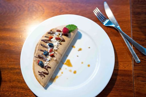 A gluten-free healthy pancake with berries and vegan chocolate sauce on a white plate with fork and spoon on a wooden table