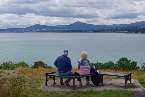 Seen from behind, an elderly couple sits on a bench in Sorrento Park, enjoying the scenic view of the bay surrounded by mountains.