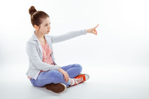 A young girl sitting cross-legged on the floor and pointing at something against a white background
