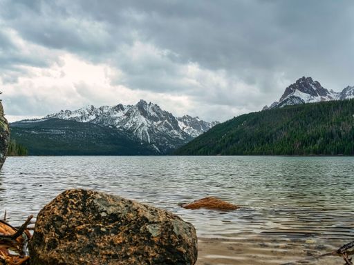 A gorgeous landscape with tall snowy peaks and forests surrounding a calm lake under a cloudy sky