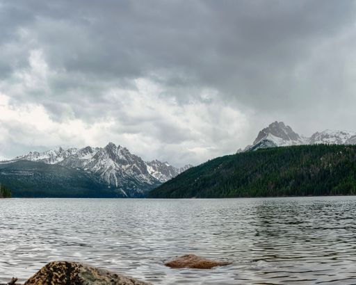 A gorgeous landscape with tall snowy peaks and forests surrounding a calm lake under a cloudy sky